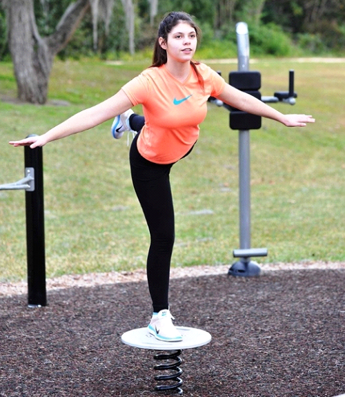 Teen performing a balance exercise on outdoor fitness equipment designed to improve stability and coordination in a park setting.